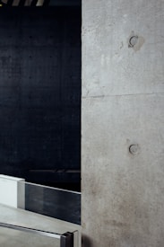 A minimalist interior space featuring a large concrete wall with visible construction markings and a metal railing in the foreground. The background is dark, adding contrast to the lighter tones of the concrete. The overall design is industrial and modern.