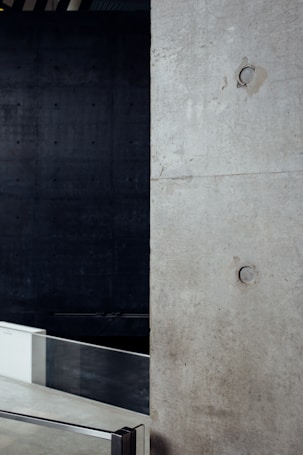 A minimalist interior space featuring a large concrete wall with visible construction markings and a metal railing in the foreground. The background is dark, adding contrast to the lighter tones of the concrete. The overall design is industrial and modern.