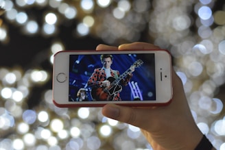 A hand is holding a smartphone displaying a performer playing a guitar on stage. The performer is wearing a vibrant, patterned jacket. The background is filled with blurred circular lights creating a bokeh effect, enhancing the atmosphere of a concert.