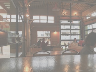 A cozy cafe with wooden interiors and large windows. Several people are seated at different tables, mingling and enjoying their time. The space is softly lit, creating a warm and inviting atmosphere. Outside, some houses and a street are visible.