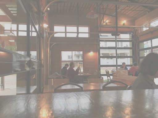 A cozy cafe with wooden interiors and large windows. Several people are seated at different tables, mingling and enjoying their time. The space is softly lit, creating a warm and inviting atmosphere. Outside, some houses and a street are visible.