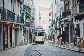 A vintage yellow tram travels along cobblestone streets lined with old buildings featuring wrought iron balconies and colorful flags. Lanterns hang from the facades, adding to the charming, historic atmosphere.