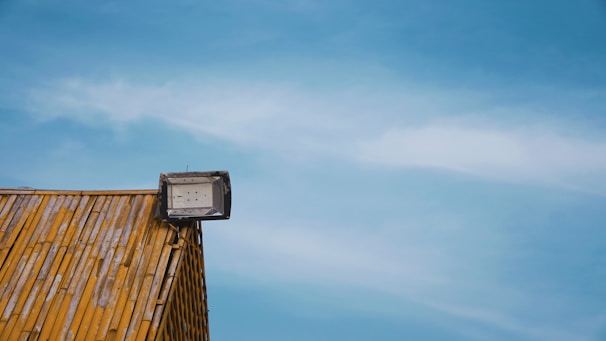 A rooftop covered with wooden shingles features a mounted floodlight against a clear blue sky.