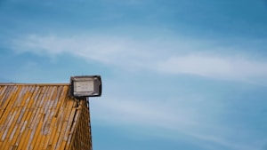 A rooftop covered with wooden shingles features a mounted floodlight against a clear blue sky.