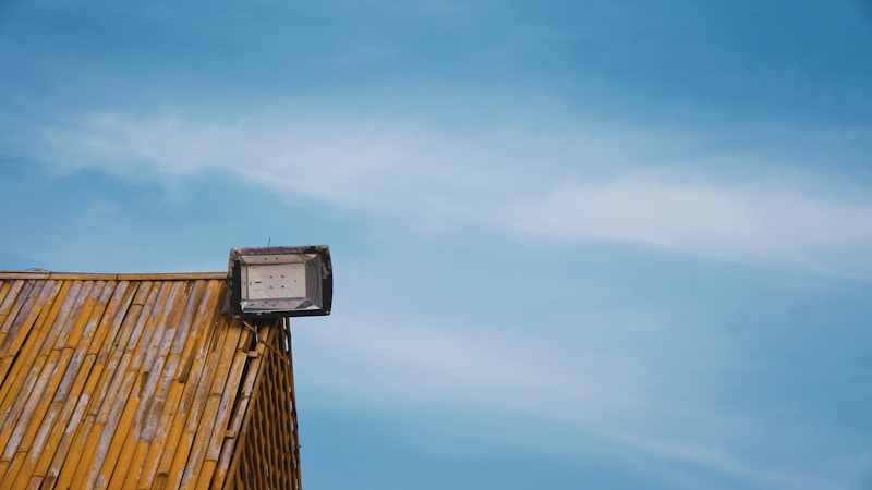 A rooftop covered with wooden shingles features a mounted floodlight against a clear blue sky.