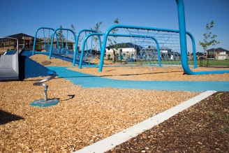 A modern playground with blue climbing structures and link bridges, set on a bed of wood chips. In the background, residential houses and small trees are visible under a clear blue sky.