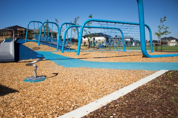 A modern playground with blue climbing structures and link bridges, set on a bed of wood chips. In the background, residential houses and small trees are visible under a clear blue sky.