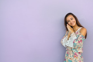 A young woman with long brown hair is leaning against a light purple wall. She is wearing a floral dress with lace details and has her hands together, slightly tilted towards her face, with a gentle smile.