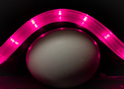 A smooth, white egg is positioned against a dark background, illuminated by a curved strip of bright pink LED lights forming an arc above it. The pink light casts a soft glow on the surface of the egg, highlighting its smooth texture and creating an interesting visual contrast with the dark surroundings.