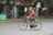 An elderly person wearing a red and gray jacket rides a bicycle along a city street. The background is blurred, indicating motion, and there are buildings and pedestrians in the distance. The bicycle has a basket in the front with what seems to be items in it.
