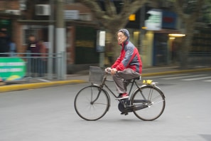 An elderly person wearing a red and gray jacket rides a bicycle along a city street. The background is blurred, indicating motion, and there are buildings and pedestrians in the distance. The bicycle has a basket in the front with what seems to be items in it.