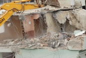 A large excavator with a yellow arm and bucket is demolishing the upper floor of a concrete building. Exposed rebar and broken concrete slabs are visible amidst the debris. Dust is rising from the destruction.