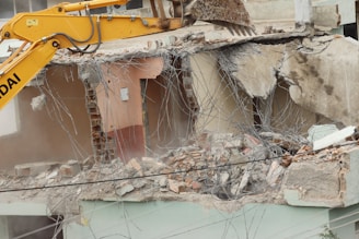 A large excavator with a yellow arm and bucket is demolishing the upper floor of a concrete building. Exposed rebar and broken concrete slabs are visible amidst the debris. Dust is rising from the destruction.