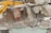 A large excavator with a yellow arm and bucket is demolishing the upper floor of a concrete building. Exposed rebar and broken concrete slabs are visible amidst the debris. Dust is rising from the destruction.
