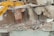 A large excavator with a yellow arm and bucket is demolishing the upper floor of a concrete building. Exposed rebar and broken concrete slabs are visible amidst the debris. Dust is rising from the destruction.