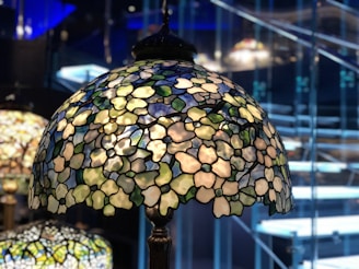 A stained glass lampshade with intricate floral patterns and colorful glass pieces is illuminated, casting soft light. The background shows blurred stairs and a modern interior setting.