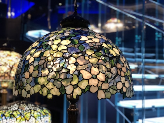 A stained glass lampshade with intricate floral patterns and colorful glass pieces is illuminated, casting soft light. The background shows blurred stairs and a modern interior setting.