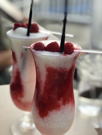 Two tall glasses filled with a creamy, iced beverage topped with fresh raspberries. The drink features layers of white and red, with raspberry garnish on top and dark straws inserted into each glass. The background is blurred, focusing attention on the refreshing drinks.