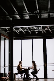 photo of two women facing each other sitting in front of table near glass wall