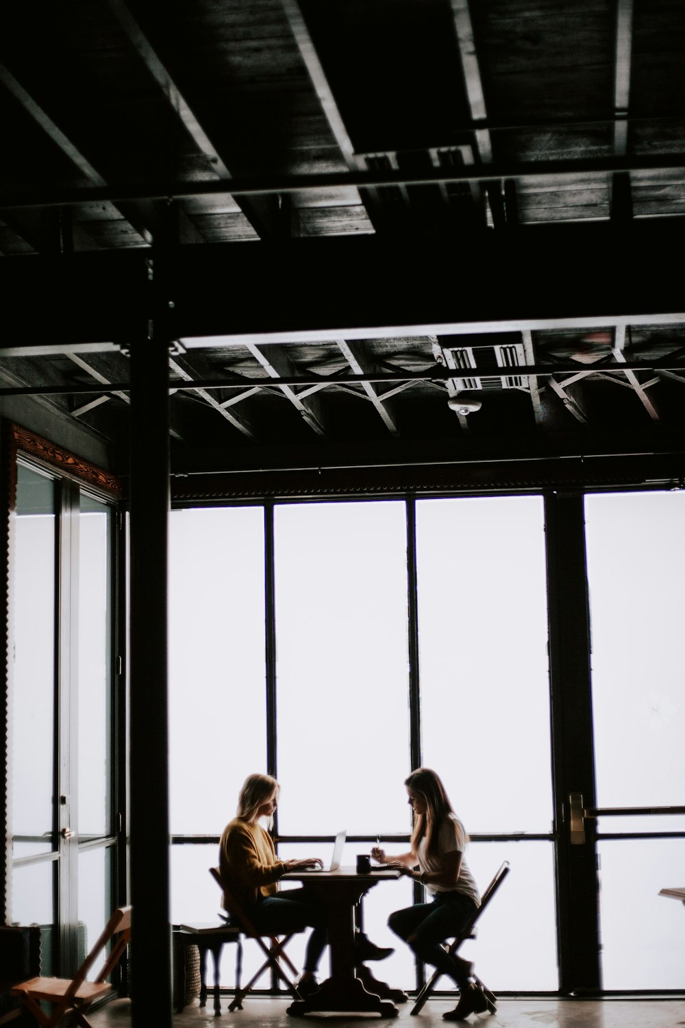photo of two women facing each other sitting in front of table near glass wall