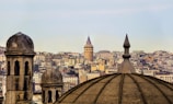 An urban landscape features the Galata Tower prominently in the background, surrounded by a dense array of buildings that form the city skyline. The foreground shows the silhouettes of traditional domes, adding a historical architectural element.