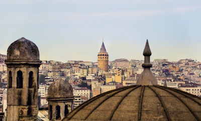 An urban landscape features the Galata Tower prominently in the background, surrounded by a dense array of buildings that form the city skyline. The foreground shows the silhouettes of traditional domes, adding a historical architectural element.