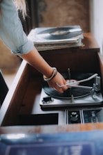 A person is adjusting the needle on a vinyl record player. There is a stack of records nearby, and the setting appears to be vintage or retro. The person's arm is adorned with bracelets, and natural lighting creates a warm atmosphere.