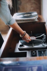 A person is adjusting the needle on a vinyl record player. There is a stack of records nearby, and the setting appears to be vintage or retro. The person's arm is adorned with bracelets, and natural lighting creates a warm atmosphere.