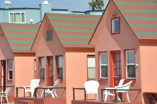 A series of quaint, peach-colored cottages with green and red striped roofs are lined up. Each cottage has a small porch with white chairs. The building behind them is painted in a contrasting teal color. A clear blue sky and a hint of palm tree are visible in the background.