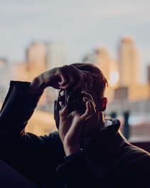 A person is holding a camera up to their face, focusing on capturing a photograph. The background features a skyline with tall buildings bathed in warm, soft lighting, possibly during sunset.