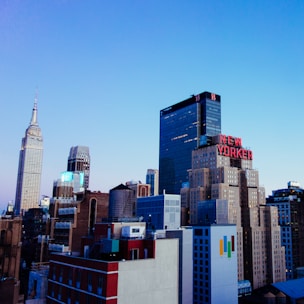 A city skyline with a prominent tall building featuring the sign 'NEW YORKER' in red letters. The scene includes several skyscrapers, with one resembling the Empire State Building. The sky is clear and blue, suggesting it is daytime.