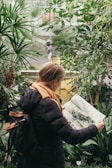 A person with a backpack, wearing a black jacket and an orange scarf, is reading a magazine or map. Surrounded by lush green plants in a dense indoor garden setting, there is a small classical statue visible in the background, adding an artistic touch to the scene.