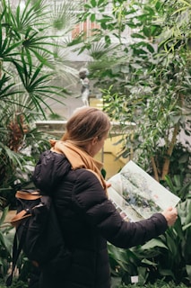A person with a backpack, wearing a black jacket and an orange scarf, is reading a magazine or map. Surrounded by lush green plants in a dense indoor garden setting, there is a small classical statue visible in the background, adding an artistic touch to the scene.