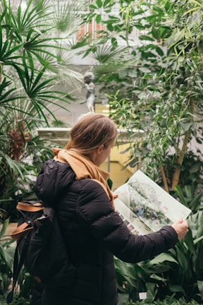 A person with a backpack, wearing a black jacket and an orange scarf, is reading a magazine or map. Surrounded by lush green plants in a dense indoor garden setting, there is a small classical statue visible in the background, adding an artistic touch to the scene.