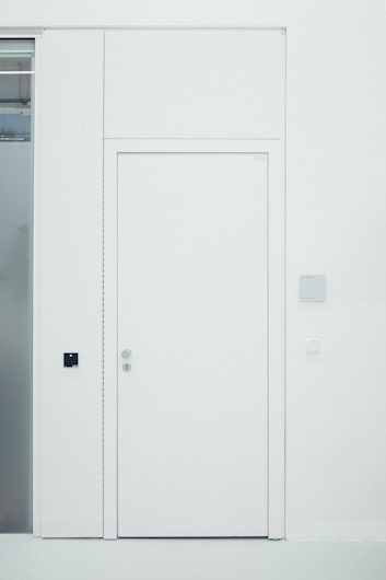 A minimalist white door is set within a white wall. The door has two metallic doorknobs, one above the other, and there is a small black square panel to the side of the door. Adjacent to the door, a frosted glass panel is visible. The design is clean and modern, with a monochromatic color scheme.