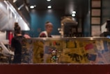 A restaurant kitchen scene with blurred people in the background, adorned with kitchen equipment. In the foreground, a golden Maneki-neko (lucky cat) figurine is prominently displayed. The background displays a deep blue wall with various framed items and a notice with Asian characters partially visible on a glass panel.