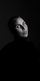 A portrait of a person with short hair, intense eyes, and a serious expression. The image is in black and white, with dramatic lighting highlighting the person's face against a dark background.