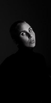 A portrait of a person with short hair, intense eyes, and a serious expression. The image is in black and white, with dramatic lighting highlighting the person's face against a dark background.