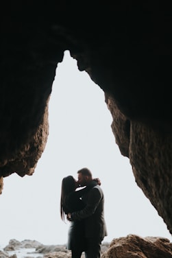 A couple embraces closely in a rocky cave opening, silhouetted against a bright sky. The scene conveys intimacy and seclusion amidst rugged natural surroundings.