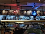 A dimly lit restaurant interior with a modern industrial design, featuring a counter displaying bright neon signs and menus. Tables and chairs are scattered throughout the dining area, and several staff members can be seen working in the background behind the counter.
