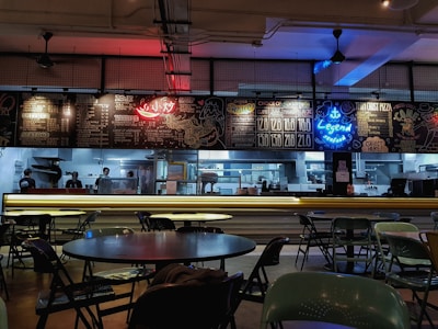 A dimly lit restaurant interior with a modern industrial design, featuring a counter displaying bright neon signs and menus. Tables and chairs are scattered throughout the dining area, and several staff members can be seen working in the background behind the counter.