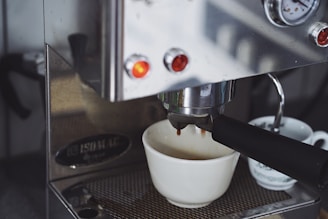 An espresso machine is brewing coffee, with a white ceramic cup positioned to catch the drip. The metal finish of the machine reflects light, and there are glowing indicator lights on the front. Next to the main cup, another smaller cup is partially visible.