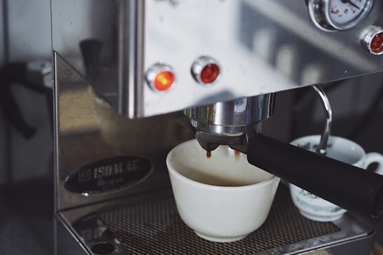 An espresso machine is brewing coffee, with a white ceramic cup positioned to catch the drip. The metal finish of the machine reflects light, and there are glowing indicator lights on the front. Next to the main cup, another smaller cup is partially visible.