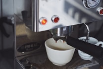 An espresso machine is brewing coffee, with a white ceramic cup positioned to catch the drip. The metal finish of the machine reflects light, and there are glowing indicator lights on the front. Next to the main cup, another smaller cup is partially visible.