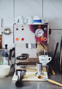 An espresso machine on a kitchen counter is surrounded by various coffee-making tools. Several cups are placed on top, and a coffee tamper and portafilter are on a cutting board. A brush and a kettle are nearby, contributing to a cozy, homey kitchen setup.