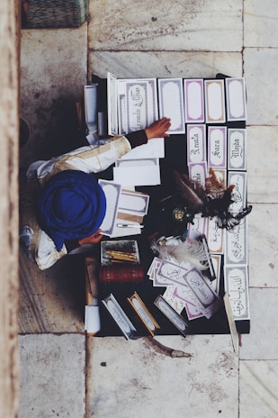 An overhead view of a person wearing a blue turban and traditional attire sitting at a table filled with ornate placards featuring calligraphic writing in different languages. The table also holds quills and art supplies, with a stone-tiled floor beneath.