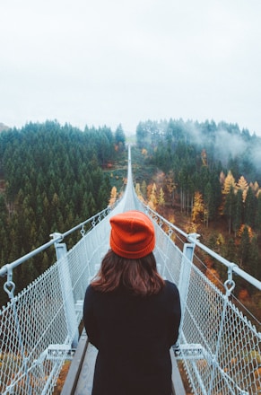 woman wearing knit cap walking on white bridge between trees during daytime
