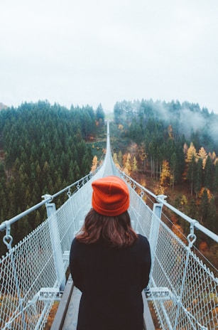 woman wearing knit cap walking on white bridge between trees during daytime