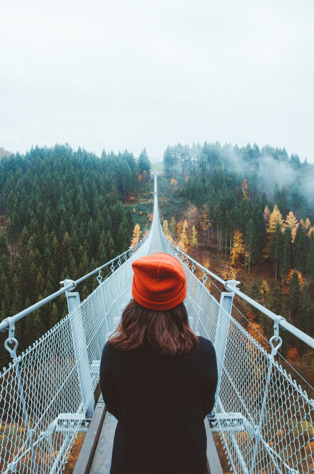 woman wearing knit cap walking on white bridge between trees during daytime