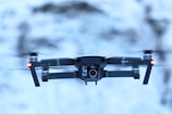 A drone is hovering in mid-air against a blurred snowy background. The drone has a sleek design with visible propellers, red lights on either side, and a camera mounted on the front.